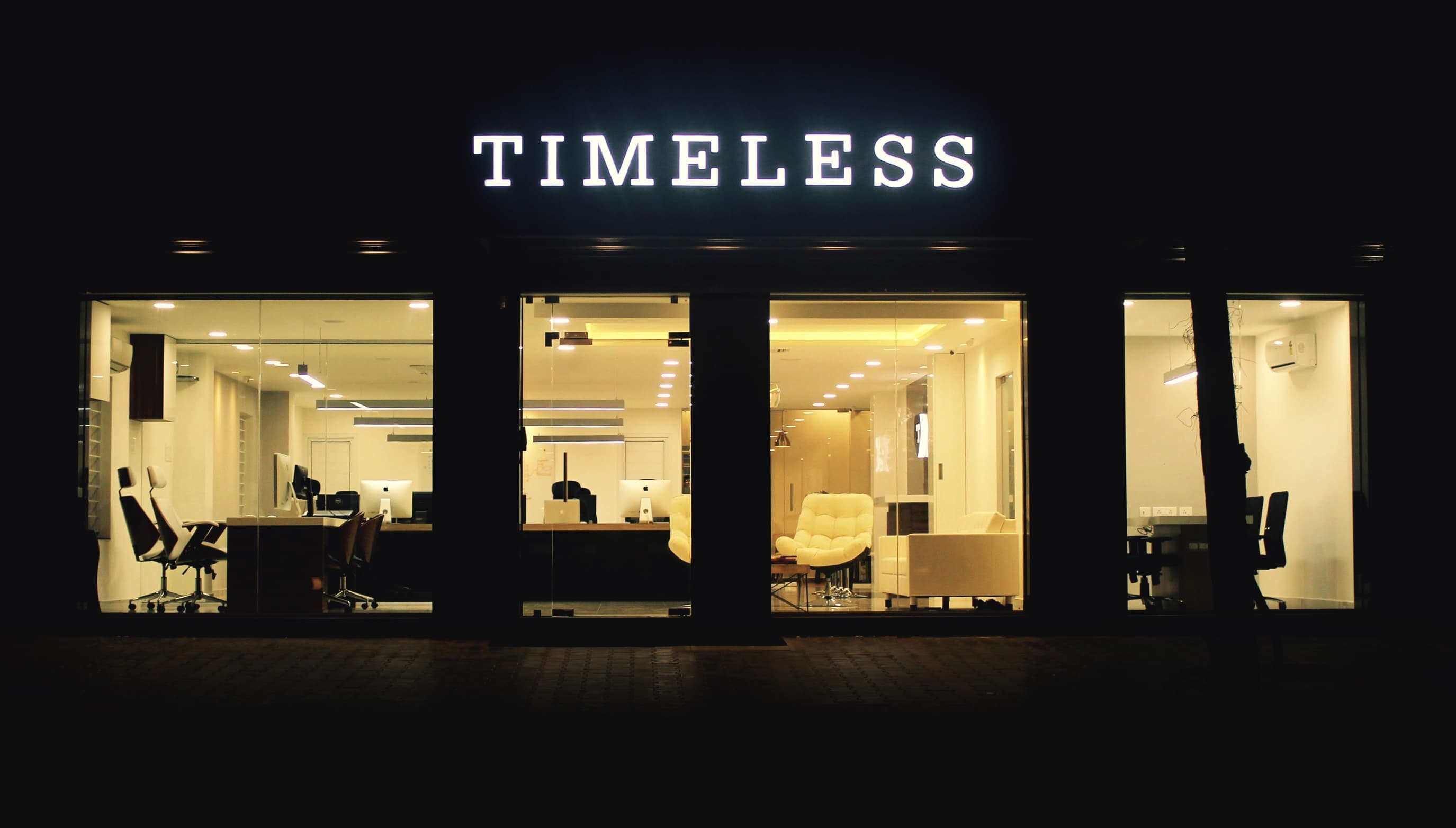 Timeless office at night with illuminated storefront displaying the company name, showcasing modern interior with workstations, lounge area, and warm lighting through floor-to-ceiling windows Timeless office at night with illuminated storefront displaying the company name, showcasing modern interior with workstations, lounge area, and warm lighting through floor-to-ceiling windows