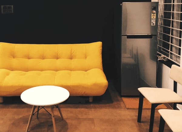Office pantry area with yellow tufted sofa, white round coffee table, and refrigerator against dark wall Office pantry area with yellow tufted sofa, white round coffee table, and refrigerator against dark wall